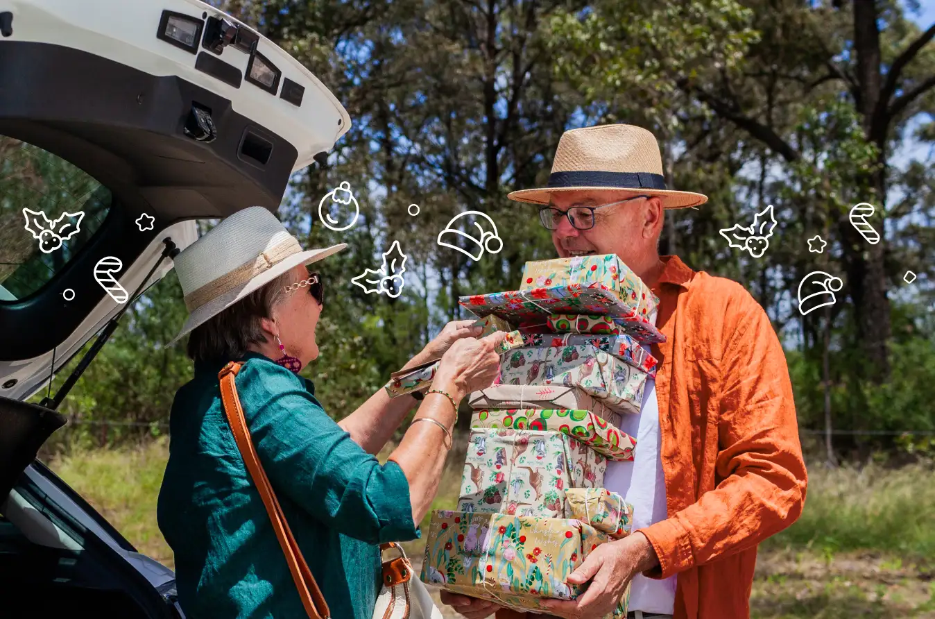 Elderly couple loading a tall stack of Christmas presents into the boot of a car.