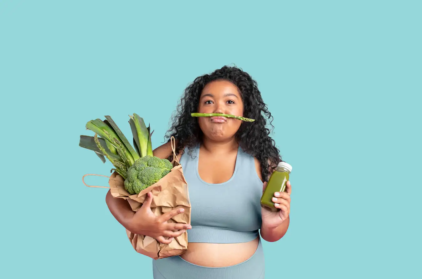 A woman posing playfully, balancing a piece of asparagus like a mustache. She is holding a brown paper bag filled with green vegetables including broccoli, leeks, and asparagus in one arm, while holding a bottle of green juice in her other hand.