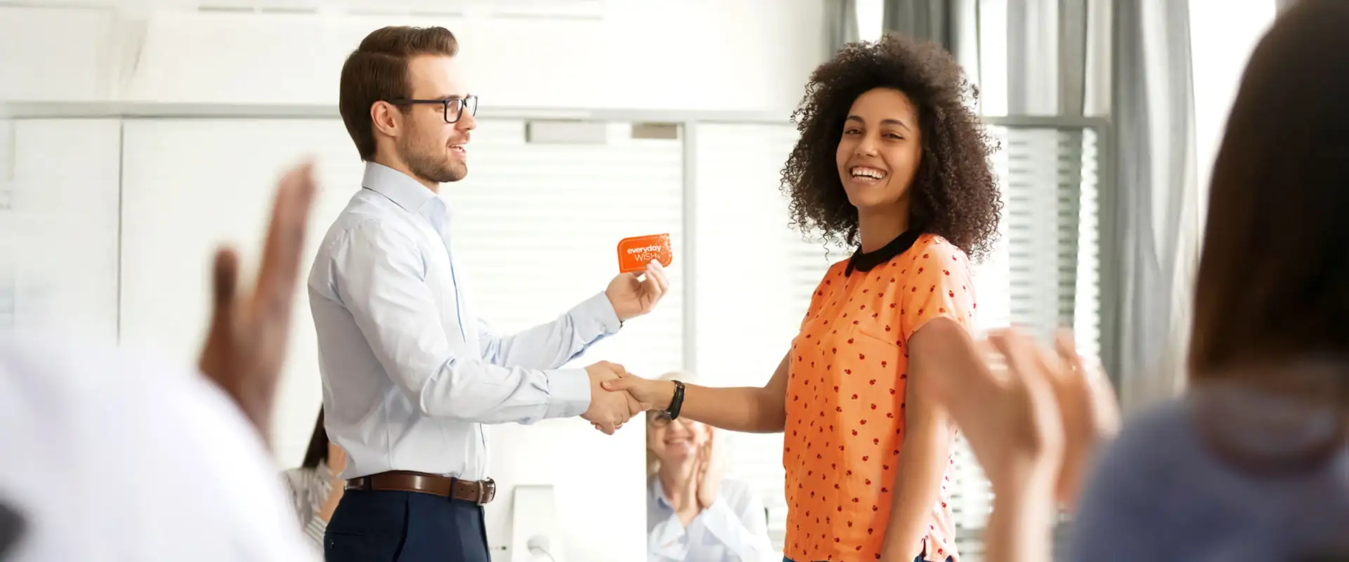 A smiling woman in a spotted orange shirt, being awarded an Everyday WISH Gift Card from a colleague while people in the background applaud.